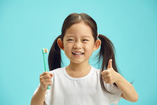 Happy and joyful korean child holding toothbrush for self teeth cleaning, observes regular oral hygiene, keep thumb up raised, cheerful smiling standing over blue isolated background. photo