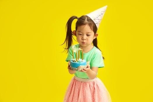 Focused Five Year Old Chinese Appearance Child in Party Hat Blow out Candles on Birthday Cake Holding it and Stands Against Yellow Background. photo