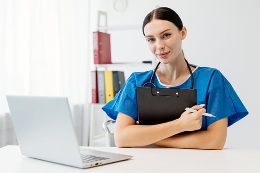 Female doctor in blue scrubs, sitting at desk with laptop and clipboard, smiling confidently while preparing for patient consultations in a bright, modern clinic environment photo