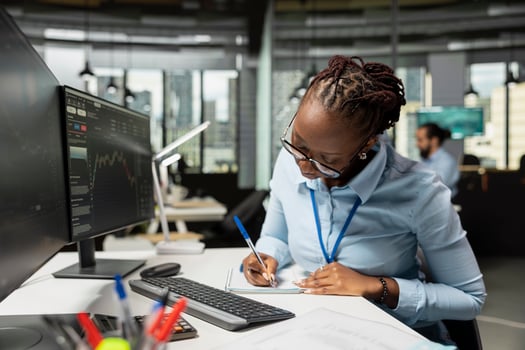 Trader in prop firm office taking notes while analyzing candlestick charts and price graphs on screen. African american woman at work writing analysis of market trends and trading strategies photo