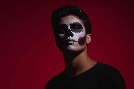 A young Hispanic male dons skeletal face paint against a crimson abyss, embodying Dia de los Muertos mystical reverie photo