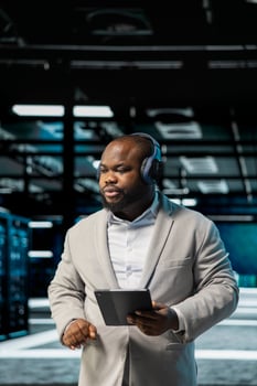 Admin listening music in data center, using tablet to oversee infrastructure mainframes. Worker wearing audio headphones walking in server farm, using tablet to supervise hardware rigs photo