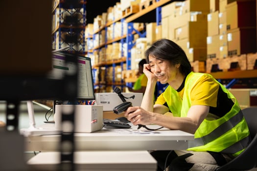 Overworked asian warehouse staff falling asleep while scanning barcodes on boxes, registering the goods in depot inventory with a scanner in hand. Fatigued worker with hi vis vest. photo