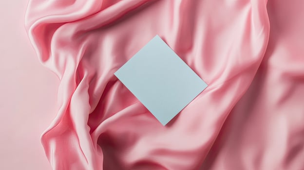 Light blue square card rests elegantly atop soft, draped light pink silk fabric photo