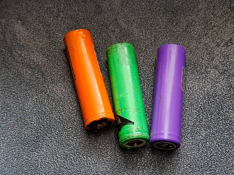 A trio of colorful cylindrical cells lies side-by-side, showcasing a vibrant orange, a lively green with visible damage, and a pristine purple, all resting on a textured surface. photo