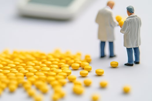 Miniature doctors assess many scattered yellow pills carefully upon a white surface in this conceptual scene photo