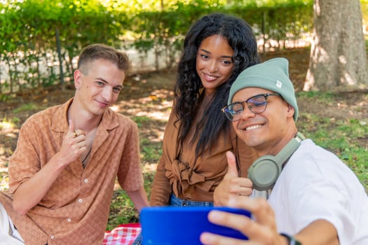 Happy multicultural friends taking selfie and showing thumbs up during picnic in park photo