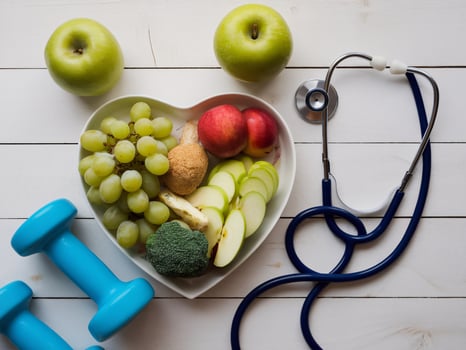 A heart-shaped bowl filled with fresh fruits and vegetables, alongside a stethoscope and dumbbells, promoting health. photo