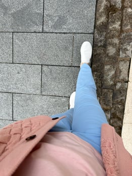 Person wearing pink sweater, light blue pants, and white shoes walking on a stone-paved sidewalk with urban texture, showcasing casual style and city life, concept of lifestyle, fashion photo