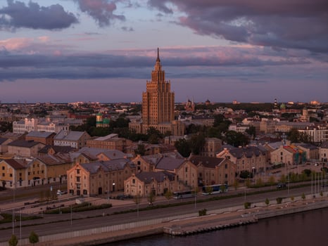 The Latvian Academy of Sciences stands at the center, surrounded by historic buildings. A waterfront area and soft pink and purple skies complete the scene. photo