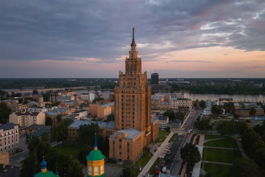 The Latvian Academy of Sciences building in Riga, Latvia, stands tall under a sunset sky, surrounded by smaller structures, greenery, and the Daugava River. photo