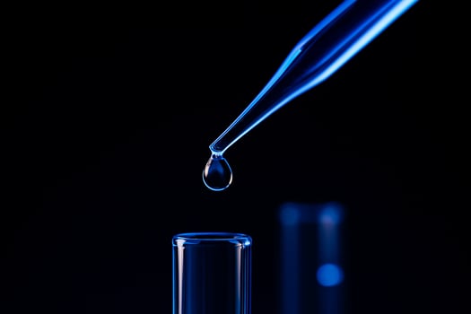 close up of a pipette releasing a clear droplet into a glass test tube against a deep blue and black background symbolizing accuracy and advanced scientific research photo