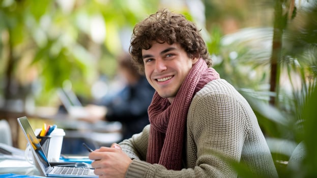 Smiling young man with curly hair in a sweater working on a laptop in a vibrant, green environment. photo