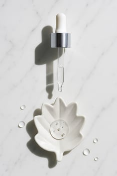 Minimal skincare scene glass dropper above a ceramic leaf dish on marble. Hard side light, crisp shadow and gel droplets. Clean, unbranded mockup for beauty wellness. photo