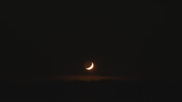 Delicate crescent moon glows above the dark horizon, with the faint outline of its full form visible against the night sky photo