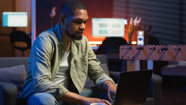 Young black adult working on his freelance tasks in the living room, adjusting his job schedule and enjoying work from his cozy home. Person navigating on web with laptop, multitasking. Camera B. photo