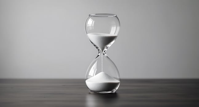 A minimalist and clean studio shot of an hourglass with white sand flowing, symbolizing the passage of time against a grey background and dark wooden surface. photo