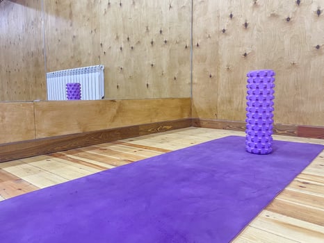 A purple yoga mat is placed on a wooden floor, accompanied by a textured purple foam roller. The room features wooden walls and a mirror reflecting the scene. photo