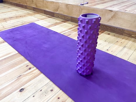 A purple foam roller stands upright on a matching purple exercise mat. The wooden floor is visible, showcasing a warm tone. Mirrors reflect the scene. photo
