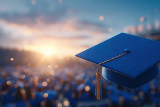 A blue graduation cap with a tassel is prominently displayed in the foreground. In the background, a crowd of graduates celebrates as the sun sets, casting a warm glow over the event. photo