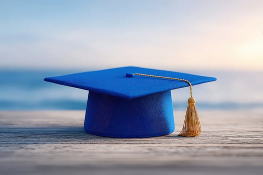 A blue graduation cap rests on a weathered wooden surface, with a golden tassel hanging to one side. The calm sea and horizon create a tranquil backdrop during twilight. photo
