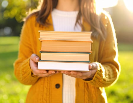 Female college student holds stack of books outdoors, wearing cozy yellow cardigan. warm sunlight creates cheerful atmosphere, perfect for studying or reading photo