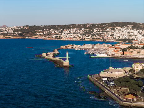 Aerial view of Chania, Crete, featuring the Venetian Harbor, iconic lighthouse, historic waterfront buildings, and surrounding hills under warm sunlight. photo