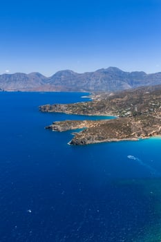 Aerial view of Voulisma Beach in Crete, Greece, featuring turquoise waters, rocky peninsulas, winding roads, scattered vegetation, and distant mountains. photo