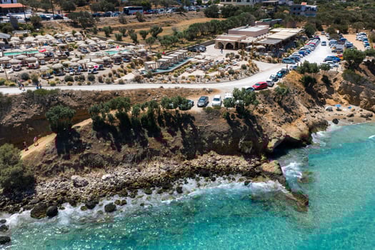 Aerial view of Voulisma Beach in Crete, Greece, showing turquoise waters, rocky shoreline, sunbeds, umbrellas, and nearby buildings along a coastal road. photo