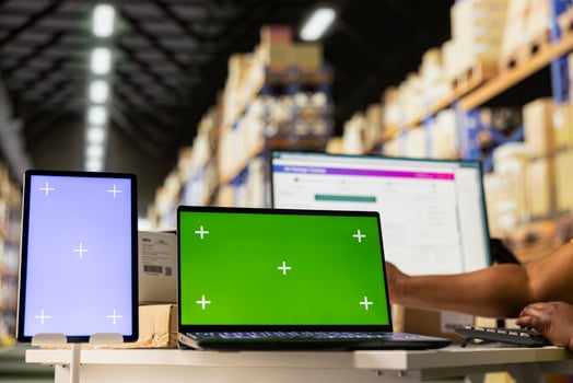 Staff reviews chroma key monitor in the stock room for data entry, inventory tracking mockups and visuals or barcode printer interfaces. Logistics worker packages cargo in the foreground. photo