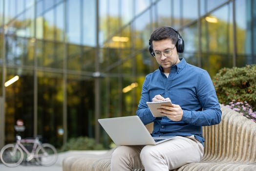Person reviewing notes while working on a laptop outdoors in a business-like setup. Background of modern architecture and a sunny environment conveying productivity and focus. photo