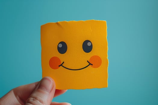 Hand holding a yellow paper with a smiling face drawn on it against a blue background. photo