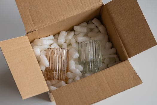Fragile glassware in pastel shades carefully packed with protective foam peanuts inside an open cardboard box. Safe packaging and shipping preparation for delicate items on a neutral background photo