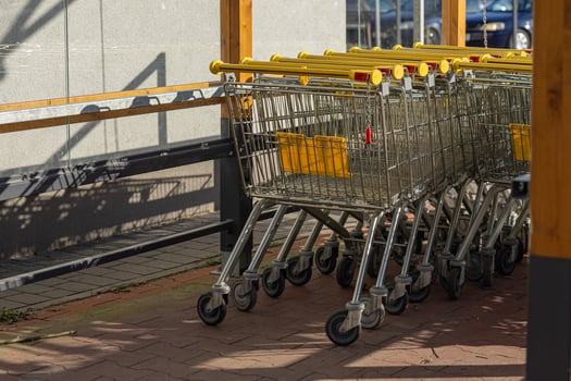 Row of metal shopping carts with yellow handles arranged in a designated cart storage area near a supermarket, illuminated by sunlight. Retail convenience, organization and shopping facilities photo