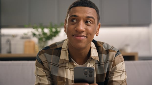 Joyful smiling African American man male guy at home using mobile phone looking for idea thinking carefree browsing smartphone application device scrolling shopping online enjoying telephone on couch photo