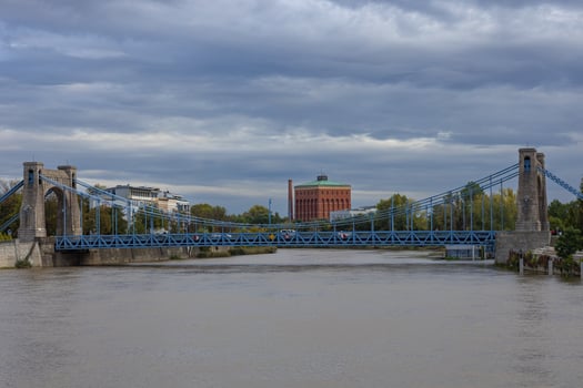 Historic blue suspension bridge over river with industrial building in background under cloudy sky on a calm day. Concept of urban architecture, transportation infrastructure, and scenic city views photo