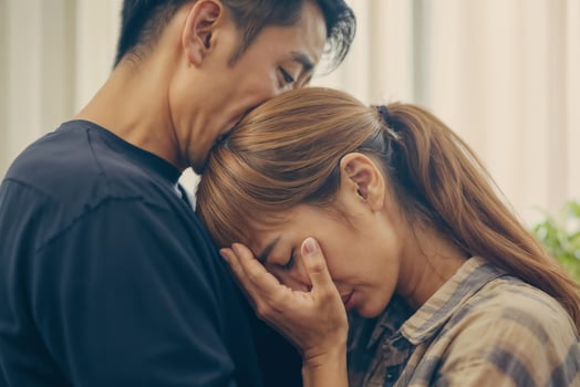 Asian man calms a woman and a woman stands close to each other, a woman cries covering her face with her hand and leans on the man's shoulder on a light blurred living room background photo