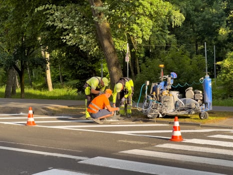 Three workers in bright safety gear are painting road lines near a tree in a green park during daytime, ensuring traffic safety photo