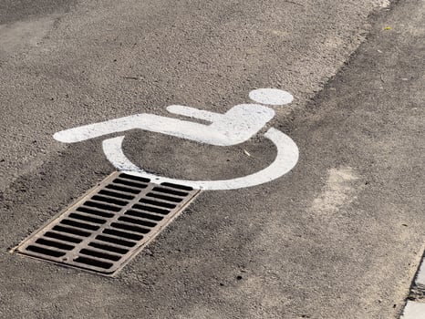 A white accessibility symbol is painted on pavement, positioned directly over a metal drainage grate in an urban setting photo