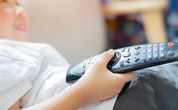 Photo of happy boy sitting on sofa at home and watching TV. Holding remote control.