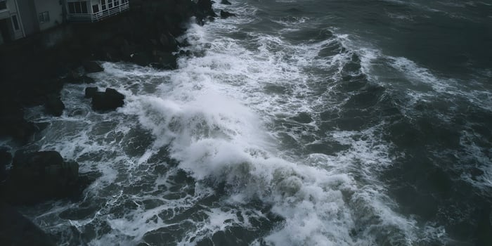 Waves crashing against rocks near a building on a cloudy day photo