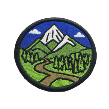 Circular embroidered patch features scenic mountain landscape with snow capped peak, green hills, and winding path through forest under blue sky with clouds png
